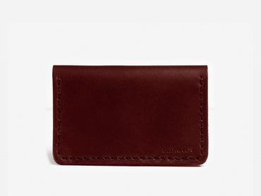 A minimalist, dark brown leather cardholder with visible stitching around the edges, displayed against a plain white background. The lower right corner has a subtle embossed logo.