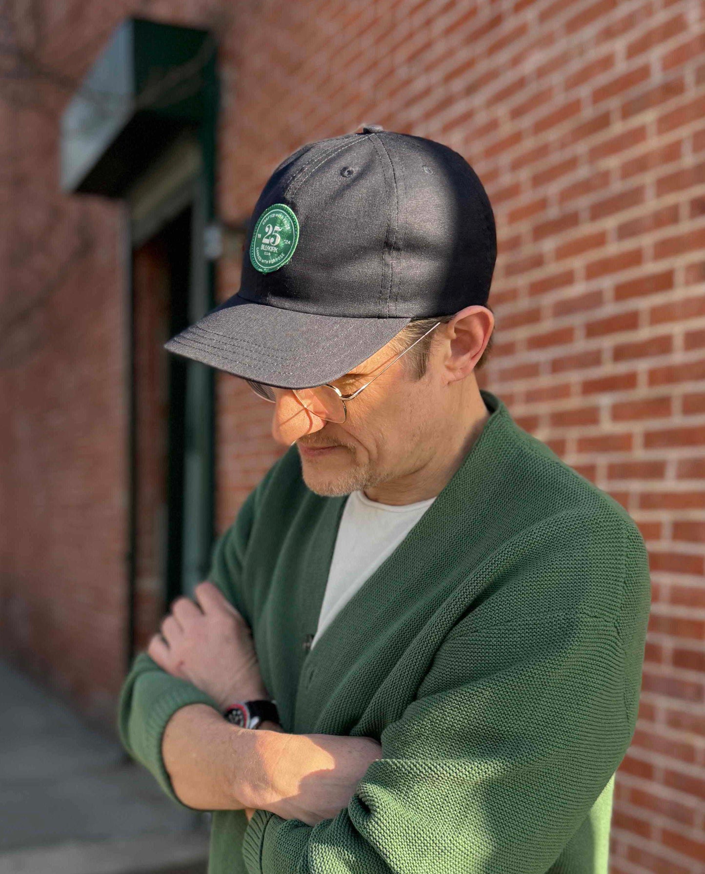 A person wearing glasses, a navy blue baseball cap, and a green cardigan stands with arms crossed in front of a brick wall, looking down.