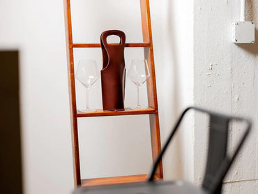 A wooden shelf holds two empty wine glasses and a brown leather wine bottle holder, set against a white wall with visible texture and a metal chair partially in the foreground.