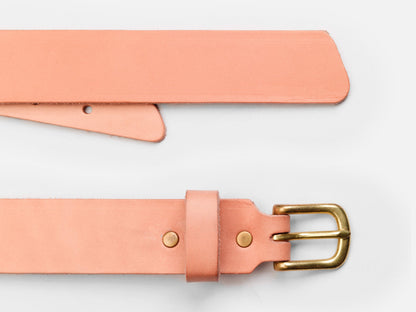 A close-up of a light pink leather belt with a brass buckle, shown laid flat on a white background. One end features the buckle and belt loop; the other end is pointed with holes for fastening.