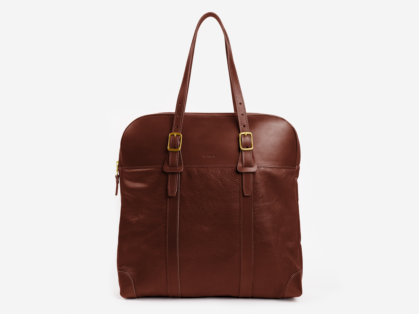 A brown leather tote bag with two shoulder straps, buckle details, a zippered top, and a subtle embossed logo on the front, set against a white background.