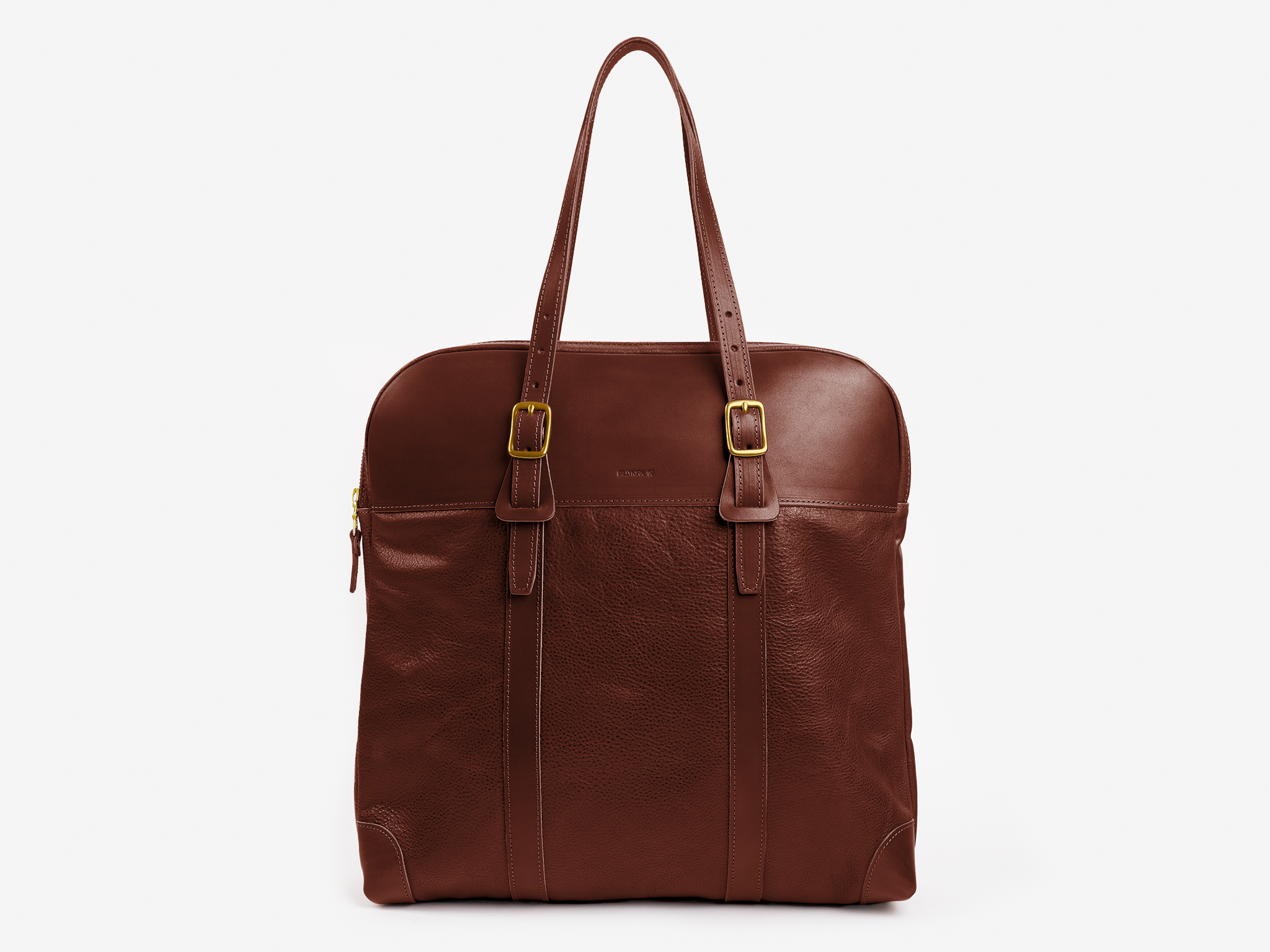 A brown leather tote bag with two shoulder straps, buckle details, a zippered top, and a subtle embossed logo on the front, set against a white background.