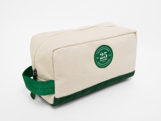 A rectangular natural canvas toiletry bag with a zipper, a green nylon webbing handle, a green suede leather base, and a green Billykirk 25 years brand patch on the front.