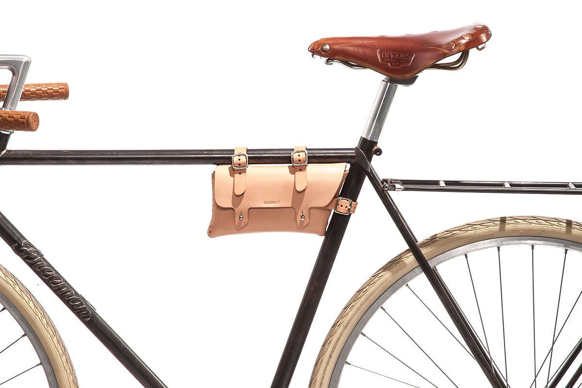 Side view of a black bicycle with tan leather seat, tan leather handlebar grips, white tires, and a small tan leather pouch attached to the frame under the seat against a white background.