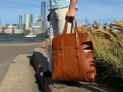 A person in striped pants holds a large brown leather bag while standing on a city sidewalk, with tall buildings and grass in the background on a sunny day.