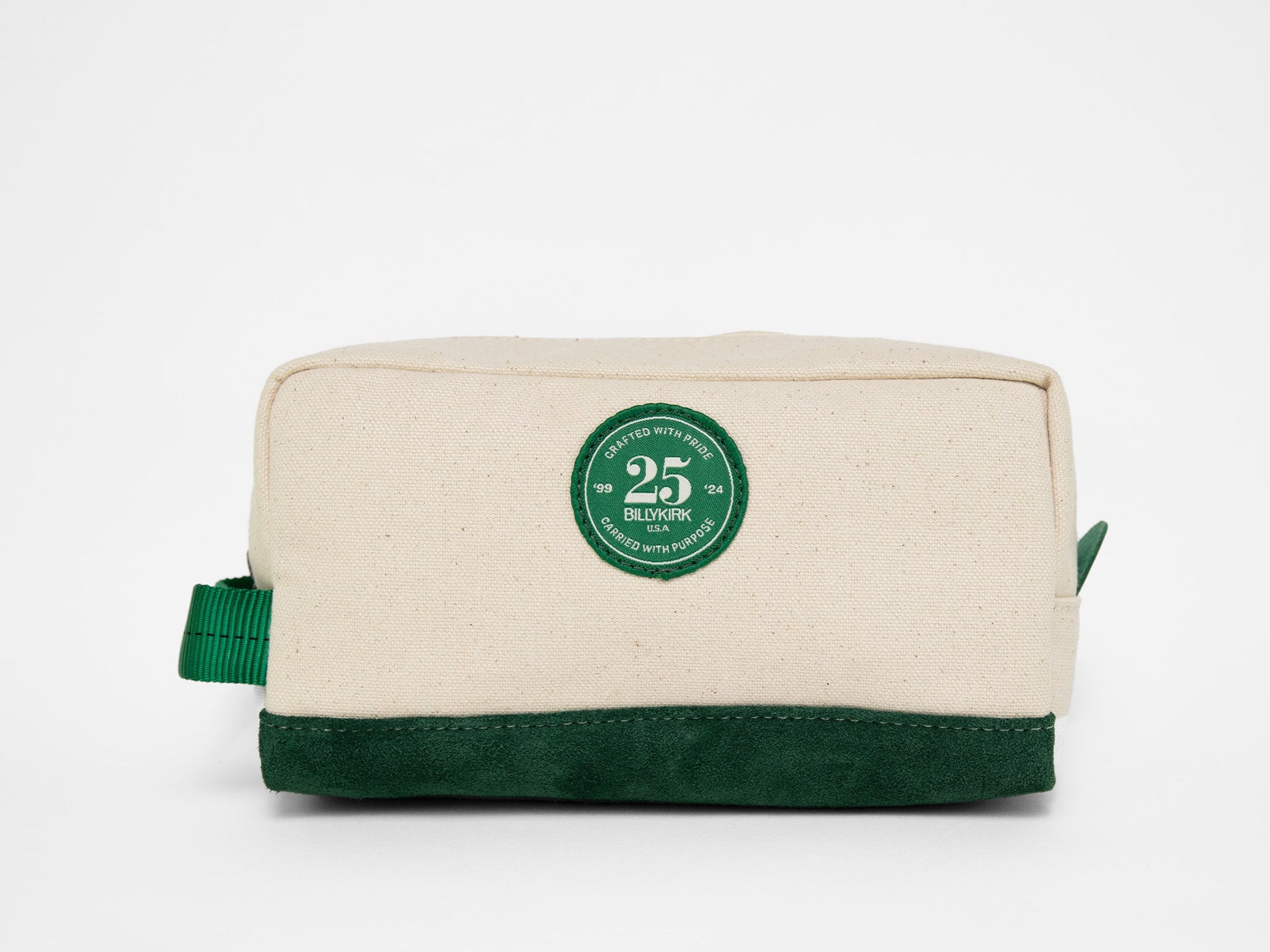 A rectangular natural canvas toiletry bag with a zipper, a green nylon webbing handle, a green suede leather base, and a green Billykirk 25 years brand patch on the front.