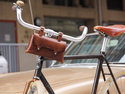 Close-up of a vintage bicycle with a brown leather saddle and matching leather handlebar bag, parked in front of a beige classic car. The focus is on the handlebar and attached bag.