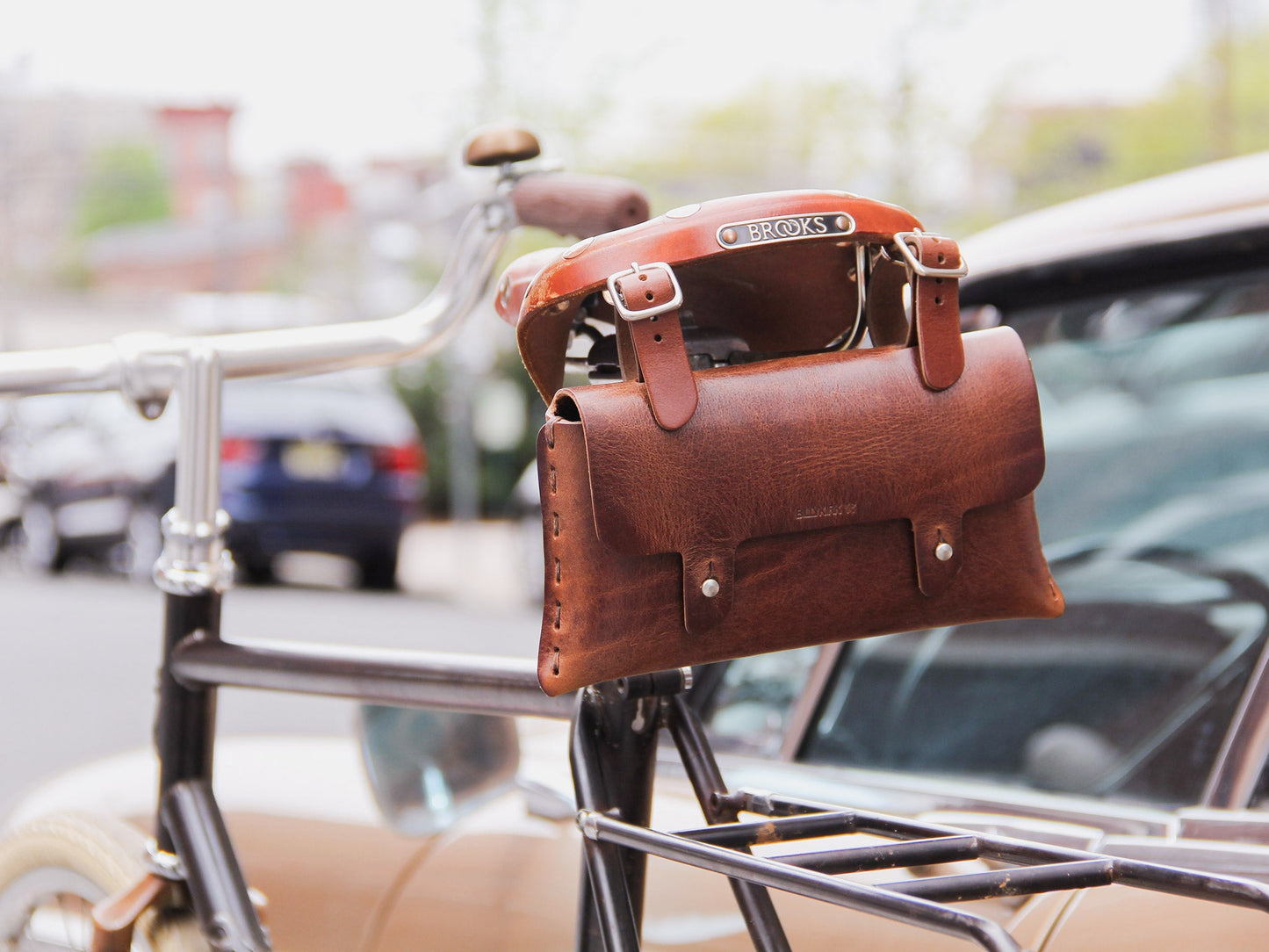 A brown leather bag is attached to the back of a bicycle seat labeled BROOKS. The bike is parked on a city street with cars and buildings blurred in the background.