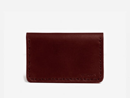 A minimalist, dark brown leather cardholder with visible stitching around the edges, displayed against a plain white background. The lower right corner has a subtle embossed logo.