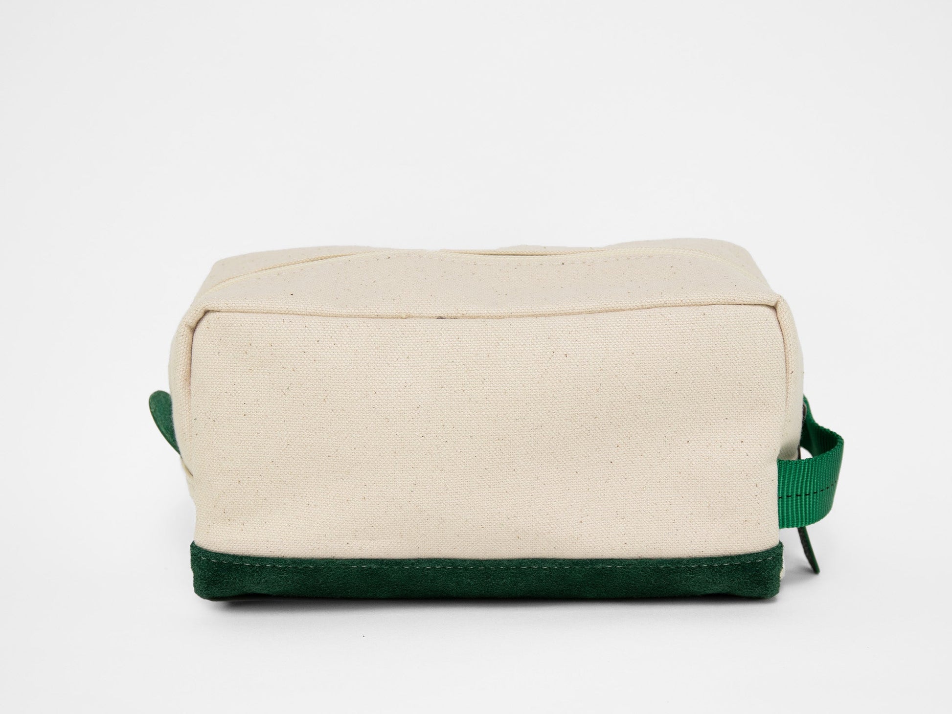 Back view of a rectangular natural canvas toiletry bag with zipper closure, green webbing handle, and suede green base.