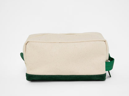 Back view of a rectangular natural canvas toiletry bag with zipper closure, green webbing handle, and suede green base.