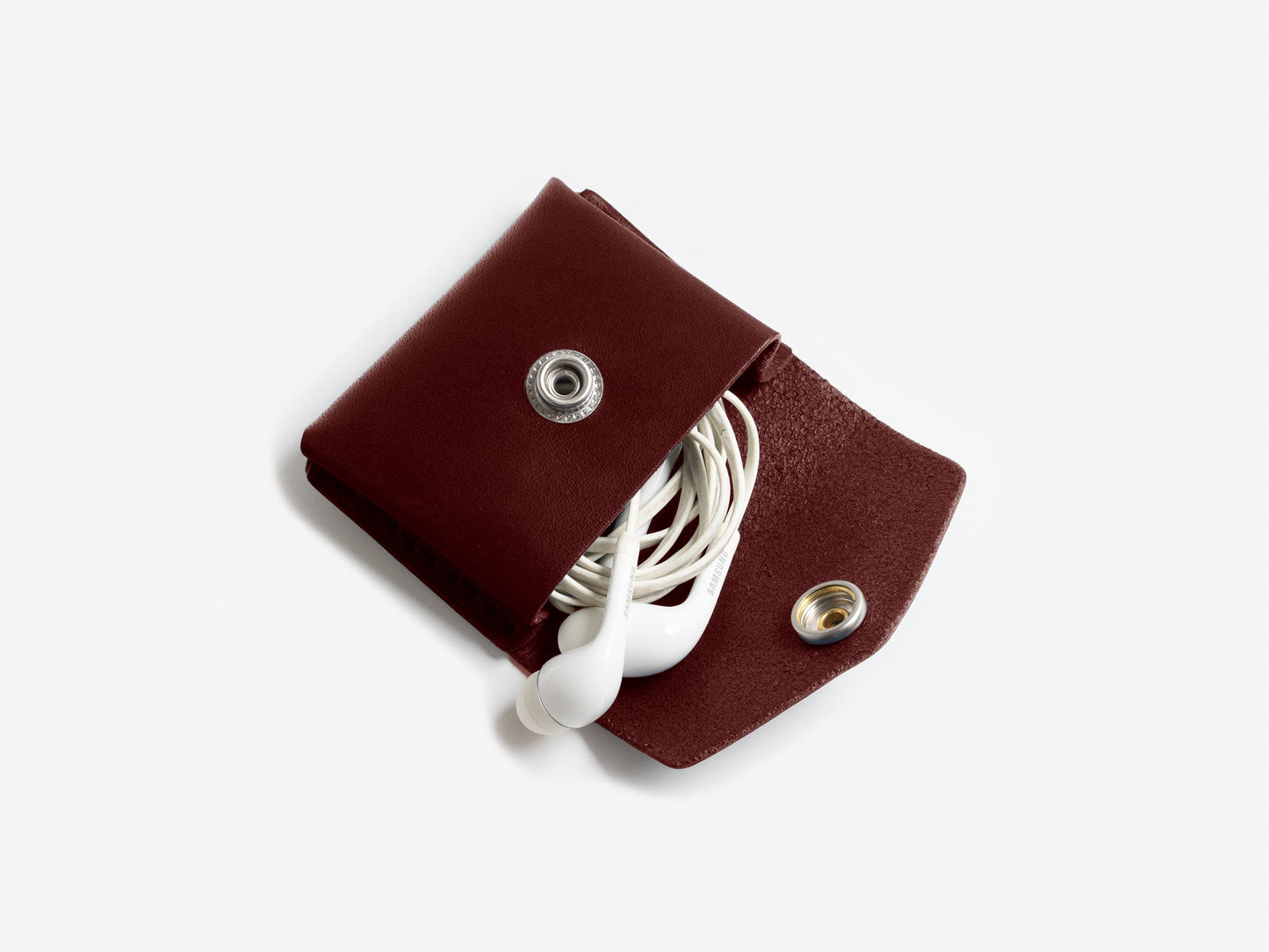 A burgundy leather coin pouch with nickel-plated snap closure, displayed with white earphones partially visible on an off-white background