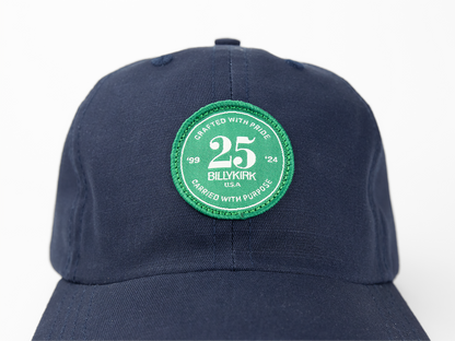 A navy blue baseball cap featuring a green and white circular patch on the front that reads “25th Billykirk” among other text.