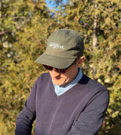 A person wearing sunglasses, an olive baseball cap, and a blue v-neck sweater stands in front of trees, looking down.