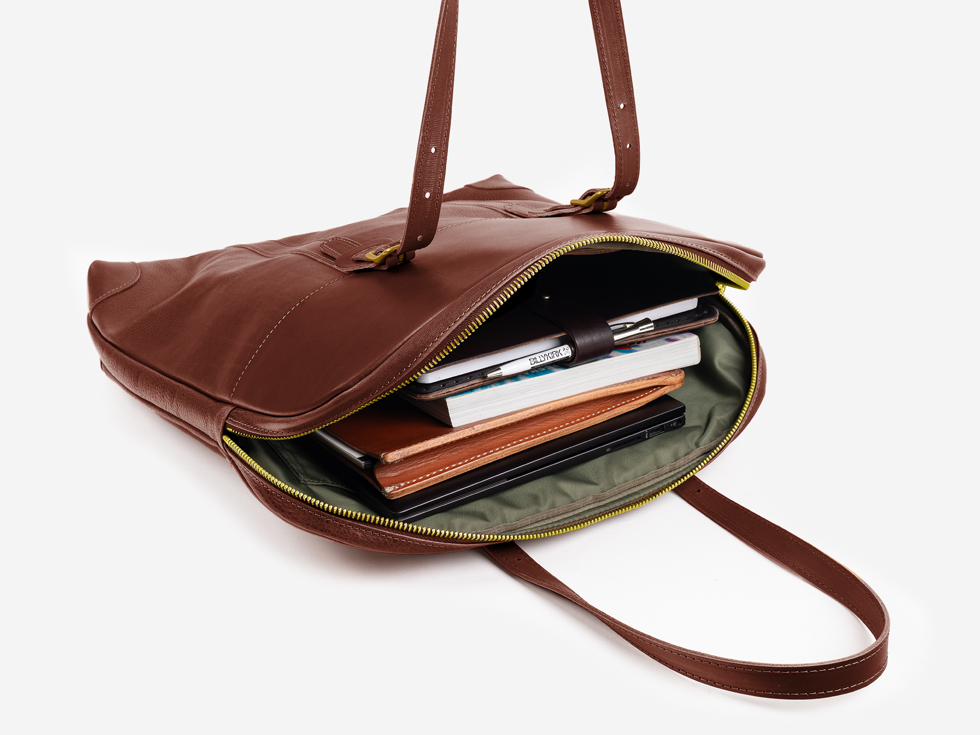A brown leather shoulder bag with a zippered opening, revealing several books, notebooks, a planner, and a pen neatly organized inside against a light background.