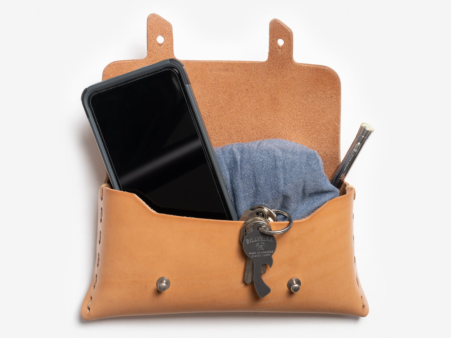 A tan leather pouch with its flap open, holding a black smartphone, a folded blue cloth, a metal pen, and a keyring with two keys. The pouch is shown against a white background.