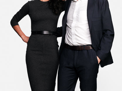 A woman in a fitted dark dress and a man in a dark suit with a white shirt stand side by side, their heads out of frame, both with one hand on their hip, against a plain white background.