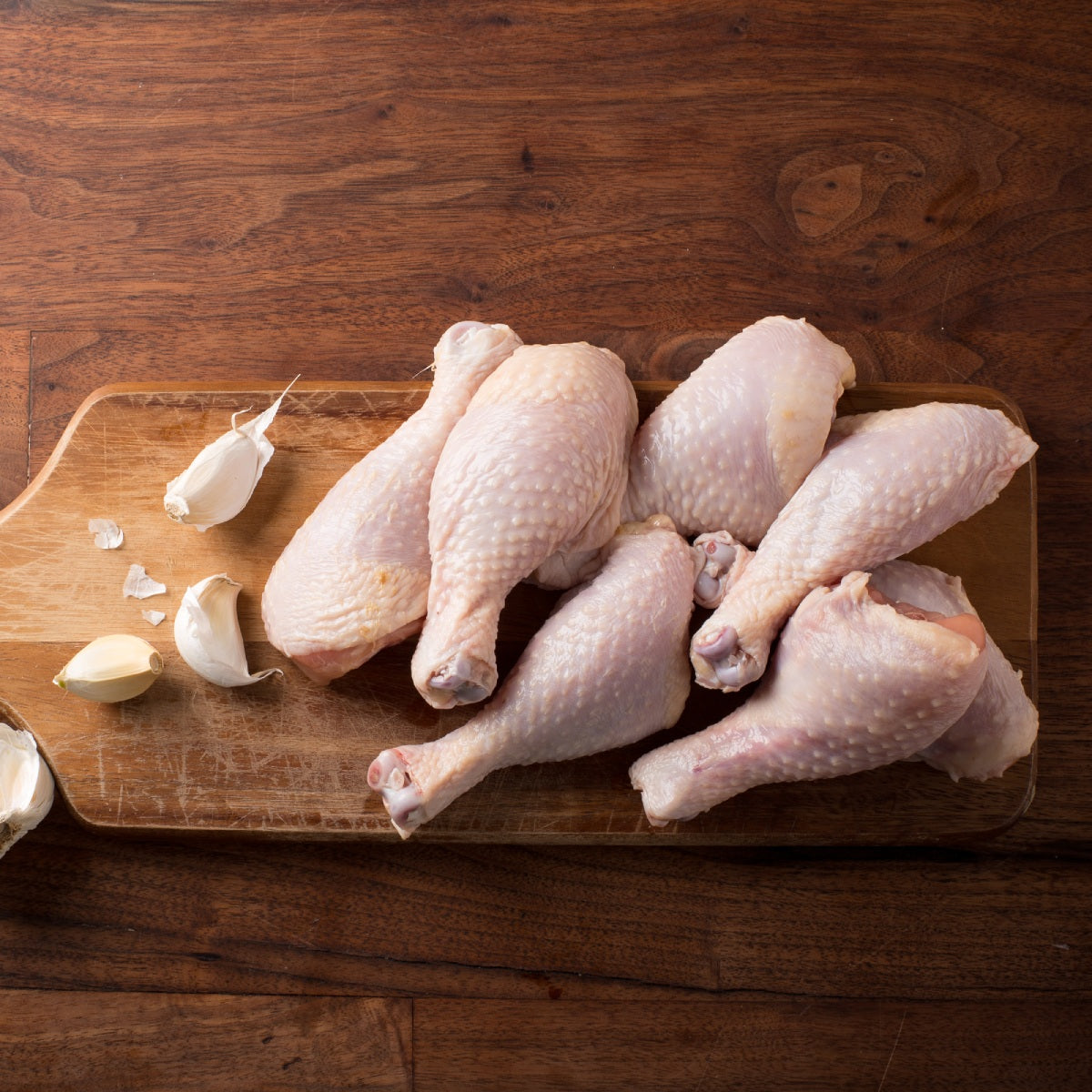 Raw pasture raised chicken drumsticks on a wooden cutting board with garlic cloves.
