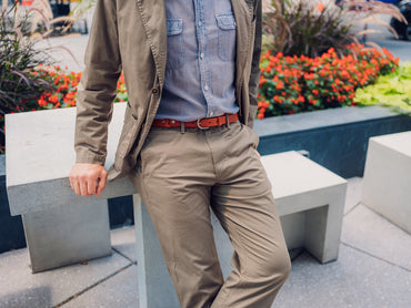 A person wearing a light blue button-up shirt, tan jacket, and khaki pants sits casually on a concrete bench near a garden with red flowers. The persons face is not visible.