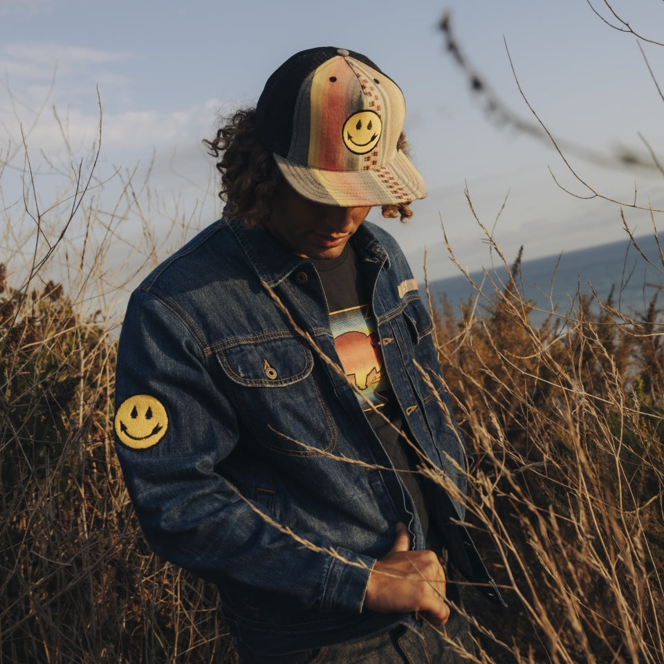 Man wearing distressed deadstock denim jacket customized by Steven Paul Judd with Native American designed patches. Wearing smiley face hat and buffalo t-shirt. 