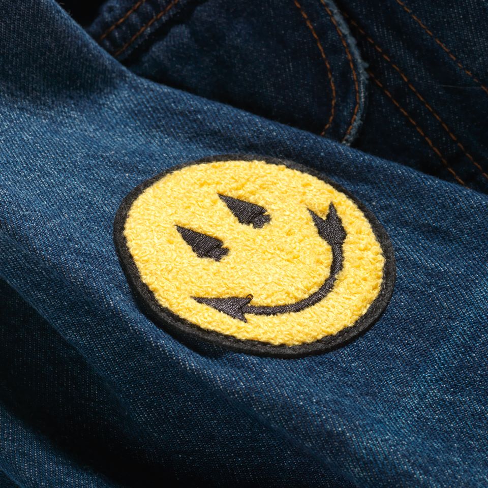 Chenille smiley face sew on patch designed by Native American Steven Paul Judd with arrowhead smile and eyes. Sewn onto a denim jacket. 