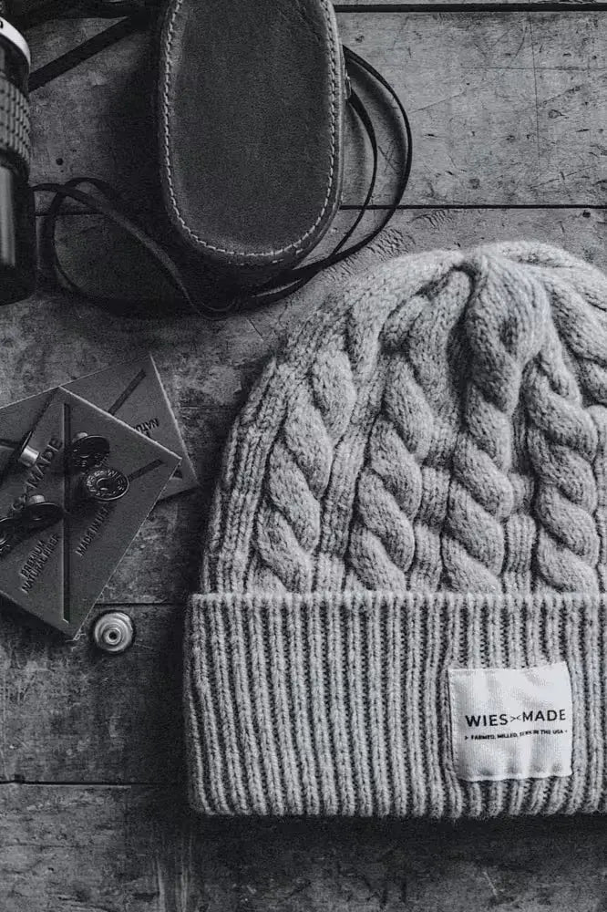Wool beanie in cable-knit design, navy color, featuring a folded cuff with a branded label, placed on a textured wooden surface alongside leather patches & a leather case