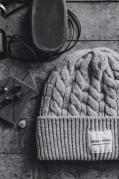 Wool beanie in cable-knit design, navy color, featuring a folded cuff with a branded label, placed on a textured wooden surface alongside leather patches & a leather case