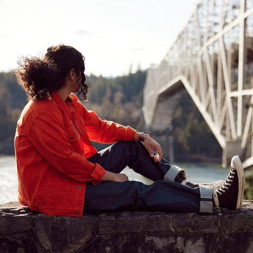 Native American wearing orange wax canvas coat made in USA by Ginew. Model outside with bridge in background.