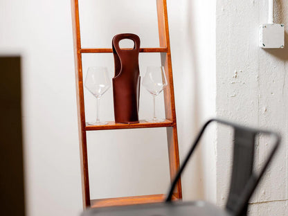 A wooden shelf holds two empty wine glasses and a brown leather wine bottle holder, set against a white wall with visible texture and a metal chair partially in the foreground.